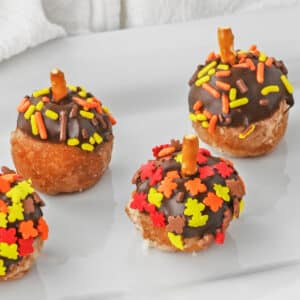 three acorn donut holes on a plate with fall sprinkles and pretzel stick as a stem.