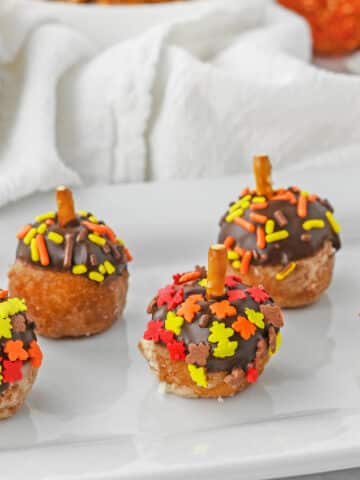 three acorn donut holes on a plate with fall sprinkles and pretzel stick as a stem.