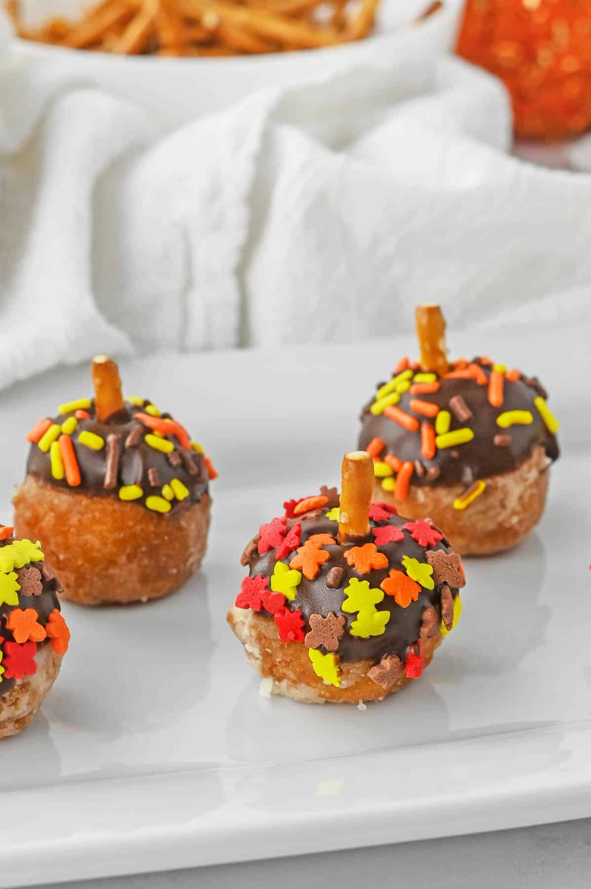 three acorn donut holes on a plate with fall sprinkles and pretzel stick as a stem.