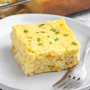 A slice of corn casserole on a white plate with a fork.