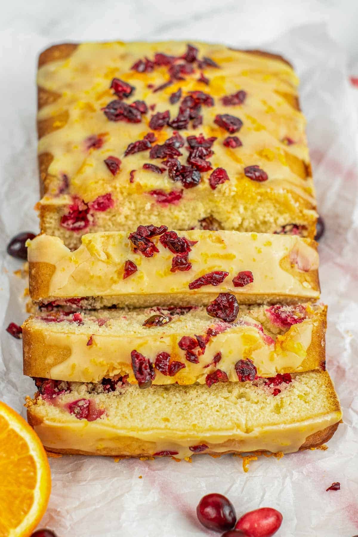 A loaf of cranberry orange bread topped with orang glaze and diced chopped cranberries with three slices cut.