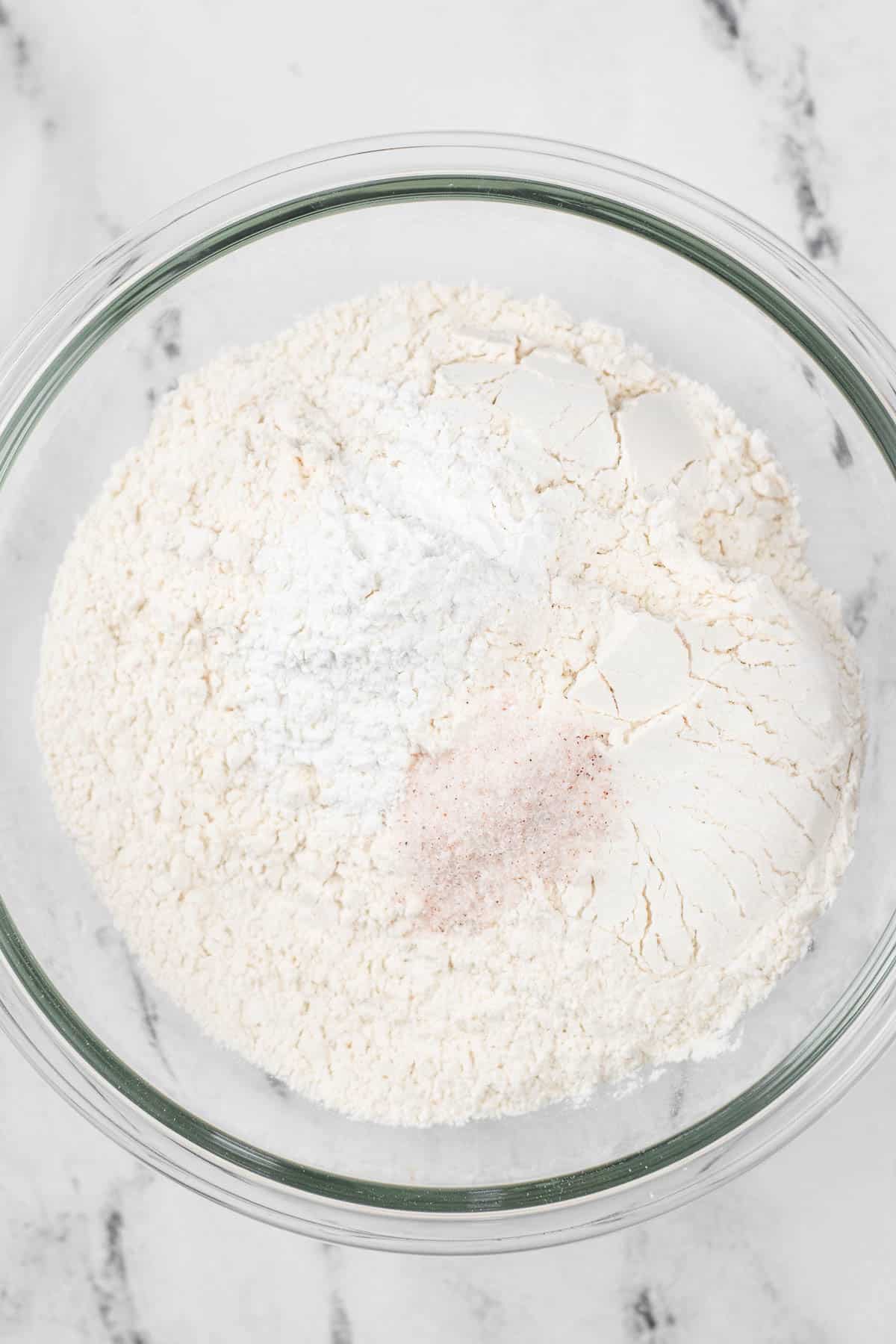 A glass mixing bowl filled with flour, with a little baking powder and salt on top.