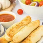 A white plate with several garlic breadsticks stacked together with a side of sauce.