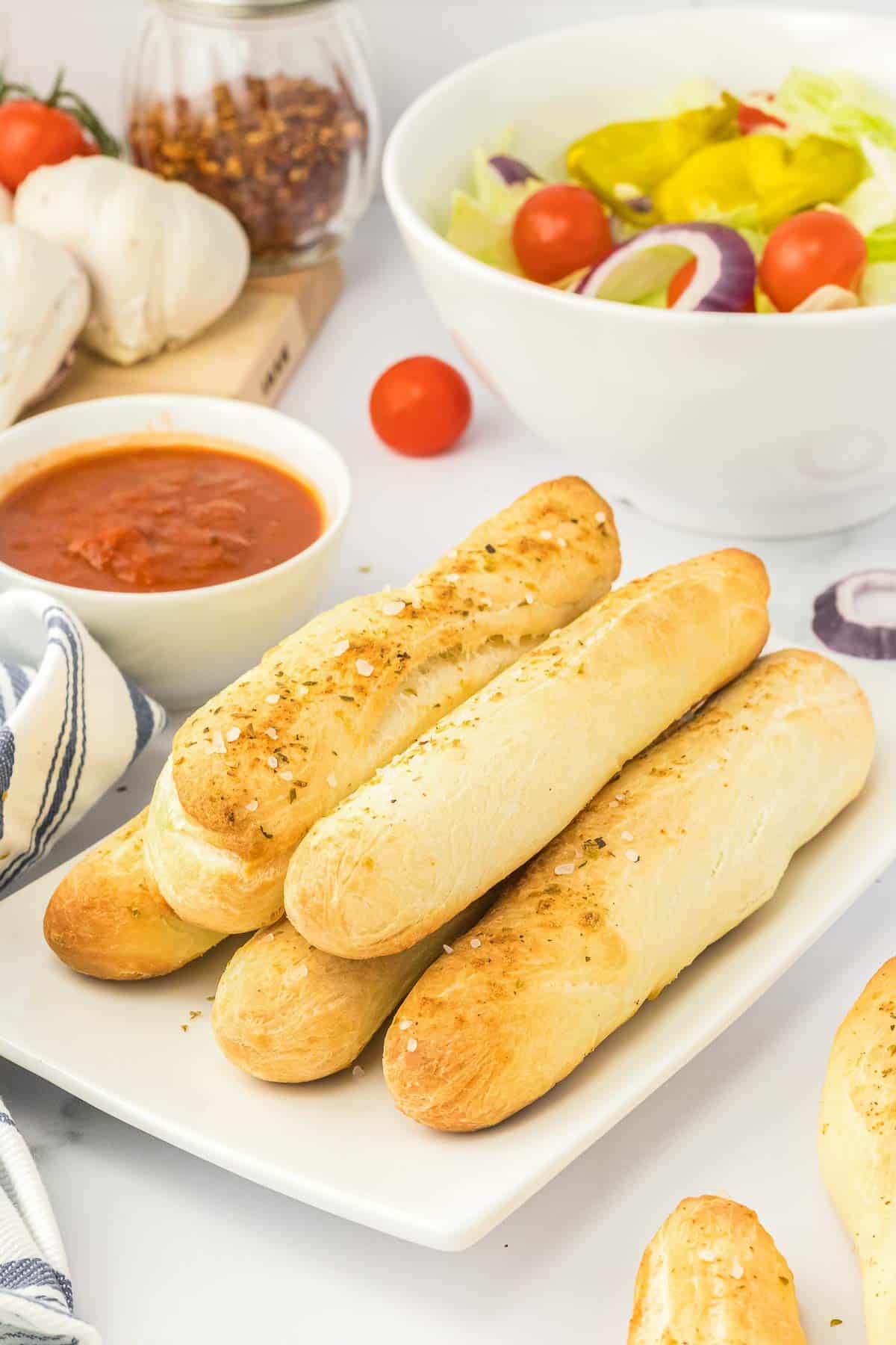 A white plate with several garlic breadsticks stacked together with a side of sauce.