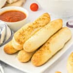 A white plate with several garlic breadsticks stacked together with a side of sauce.