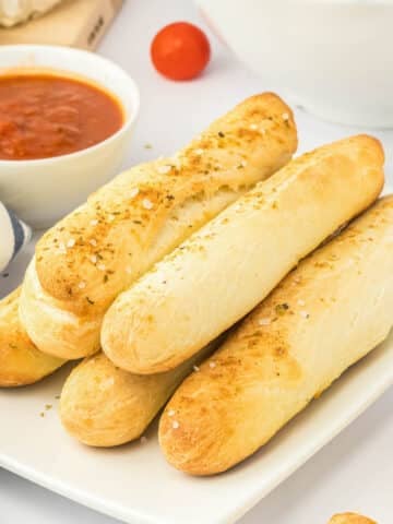 A white plate with several garlic breadsticks stacked together with a side of sauce.