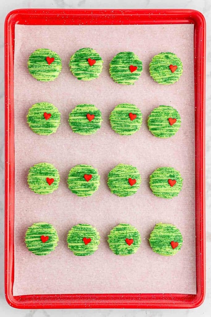 A baking sheet of grinch oreos with candy melt drizzle, green sugar and red candy hearts on them.