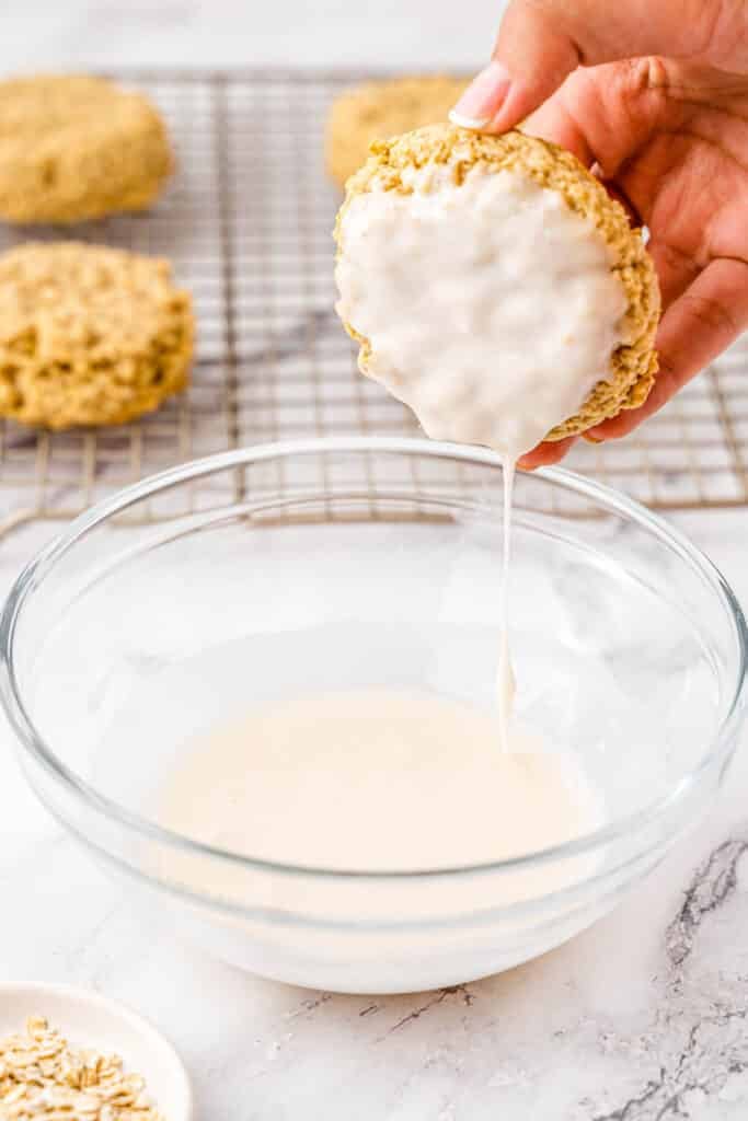 Icing on oatmeal cookie dripping into bowl held by woman's hand.