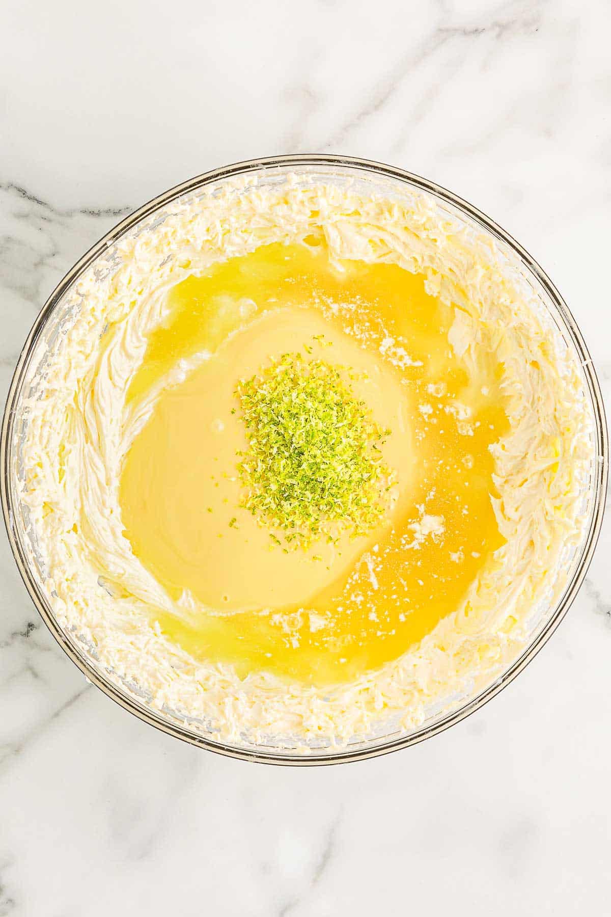 Condensed milk, lime juice, and zest added into whipped cream in a large glass mixing bowl.