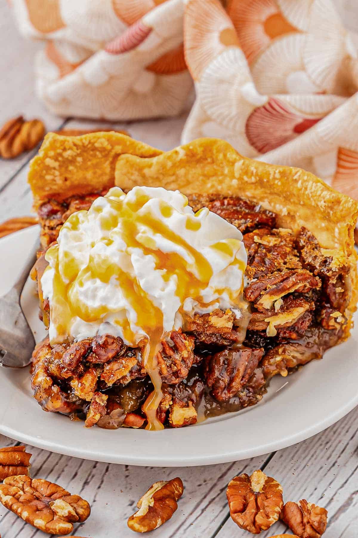 A slice of pecan pie on a white plate topped with whipped cream a caramel drizzle.