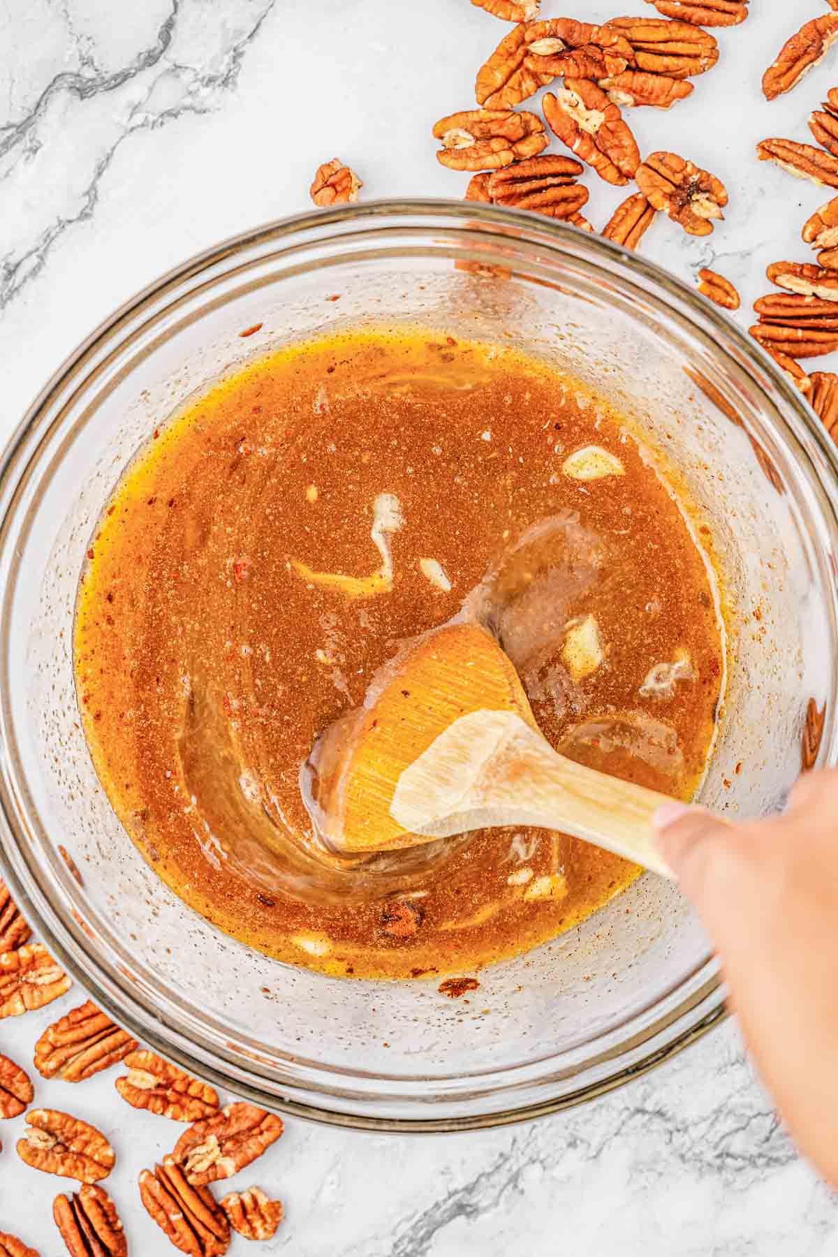 A person stirring a caramel like mixture in a glass bowl with a wooden spoon.