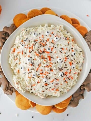 A bowl of funfetti dip topped with orange and chocolate sprinkles and surrounded by chocolate animal crackers and vanilla wafers.