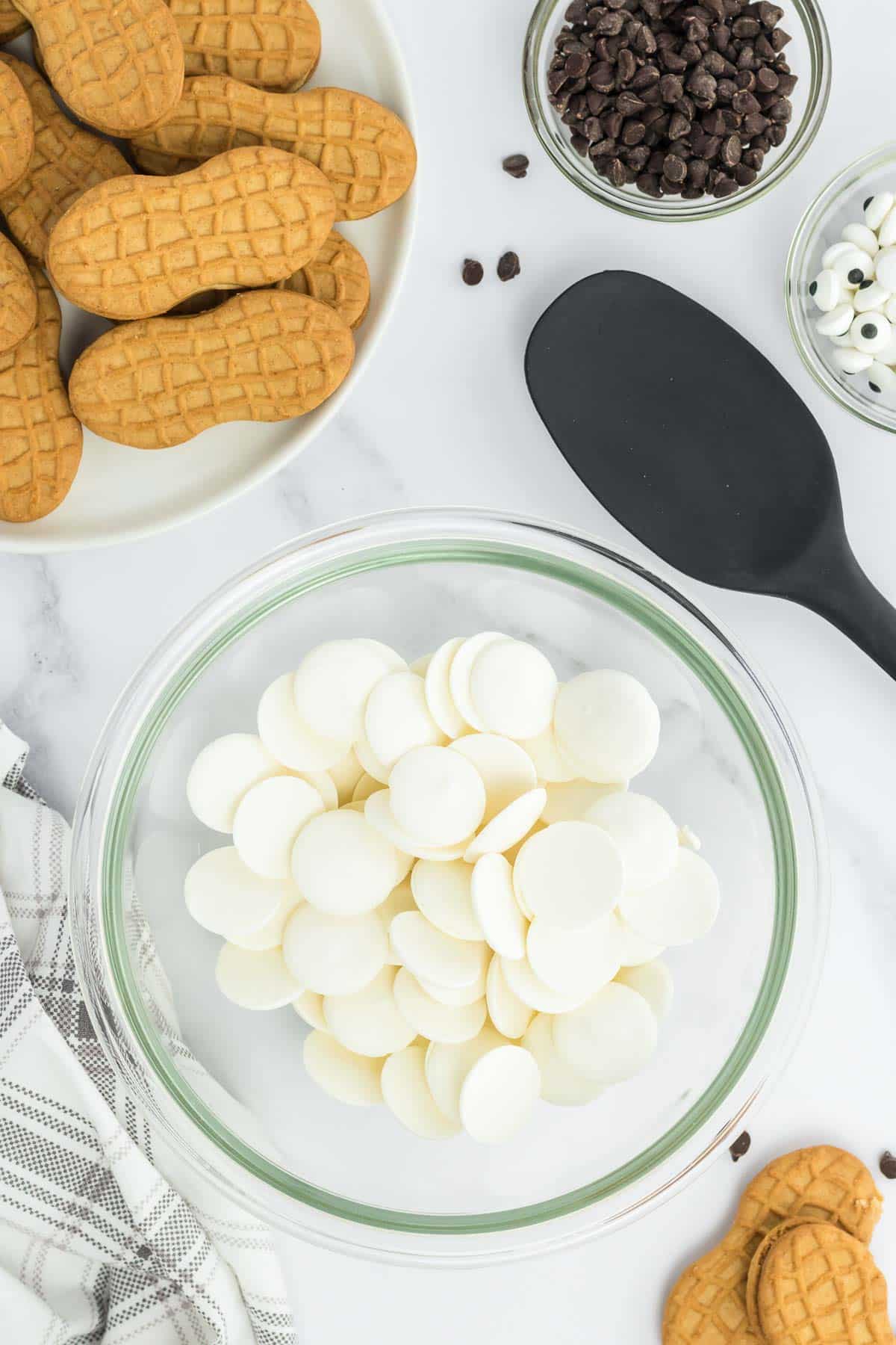 A bowl of white candy melts in a glass mixing bowl surrounded by peanut butter cookies, chocolate chips and a spoon on a marble counter.