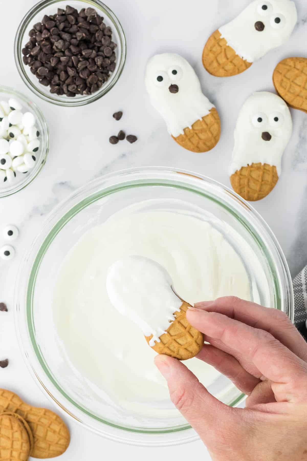 A person is dipping a nutter butter cookie into white icing in a glass bowl.