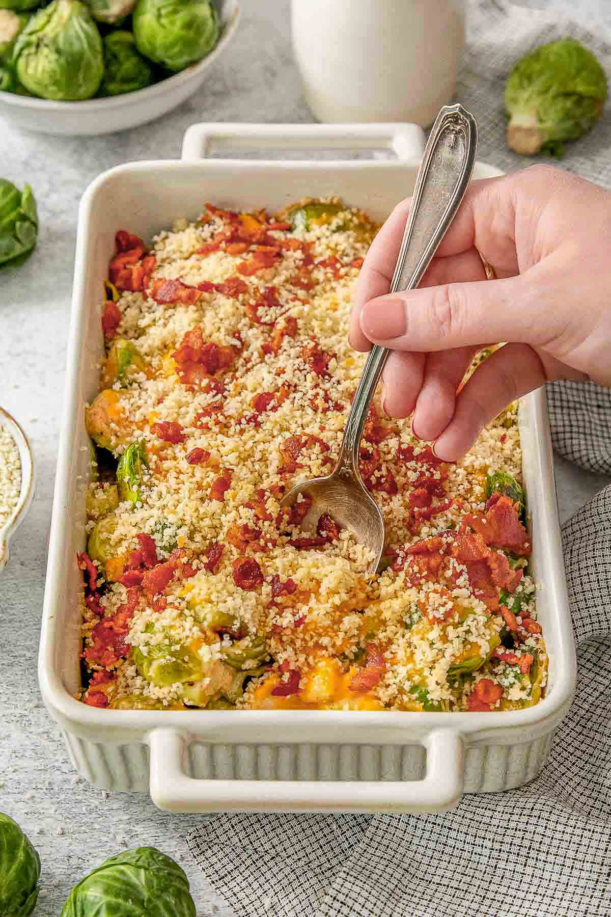 Brussels sprouts in a casserole dish with a spoon.Brussels sprout casserole in a large white baking dish topped with breadcrumbs with woman holding a spoon inserted into dish.