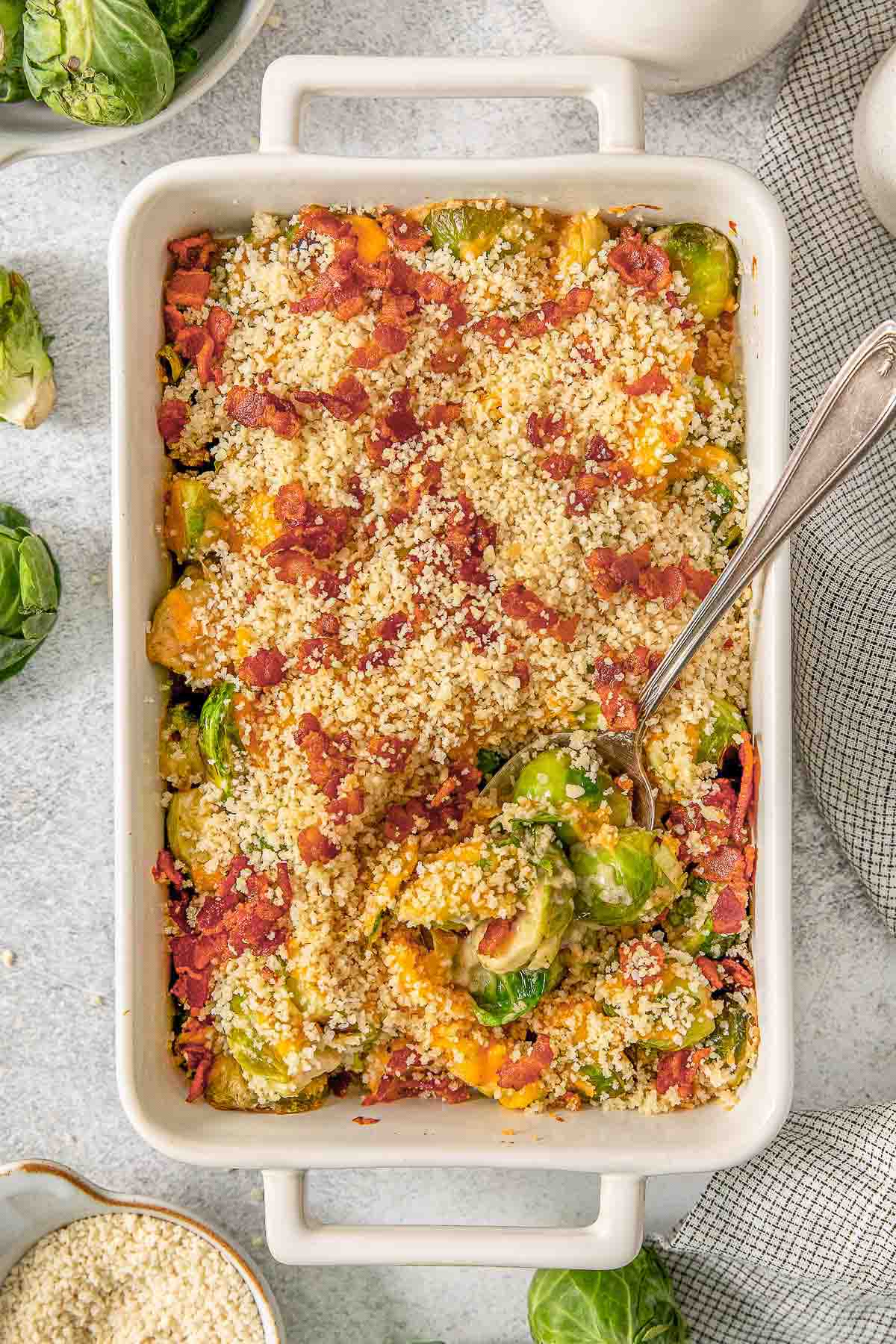 Brussels sprout casserole in a large white baking dish topped with breadcrumbs with a spoon inserted.