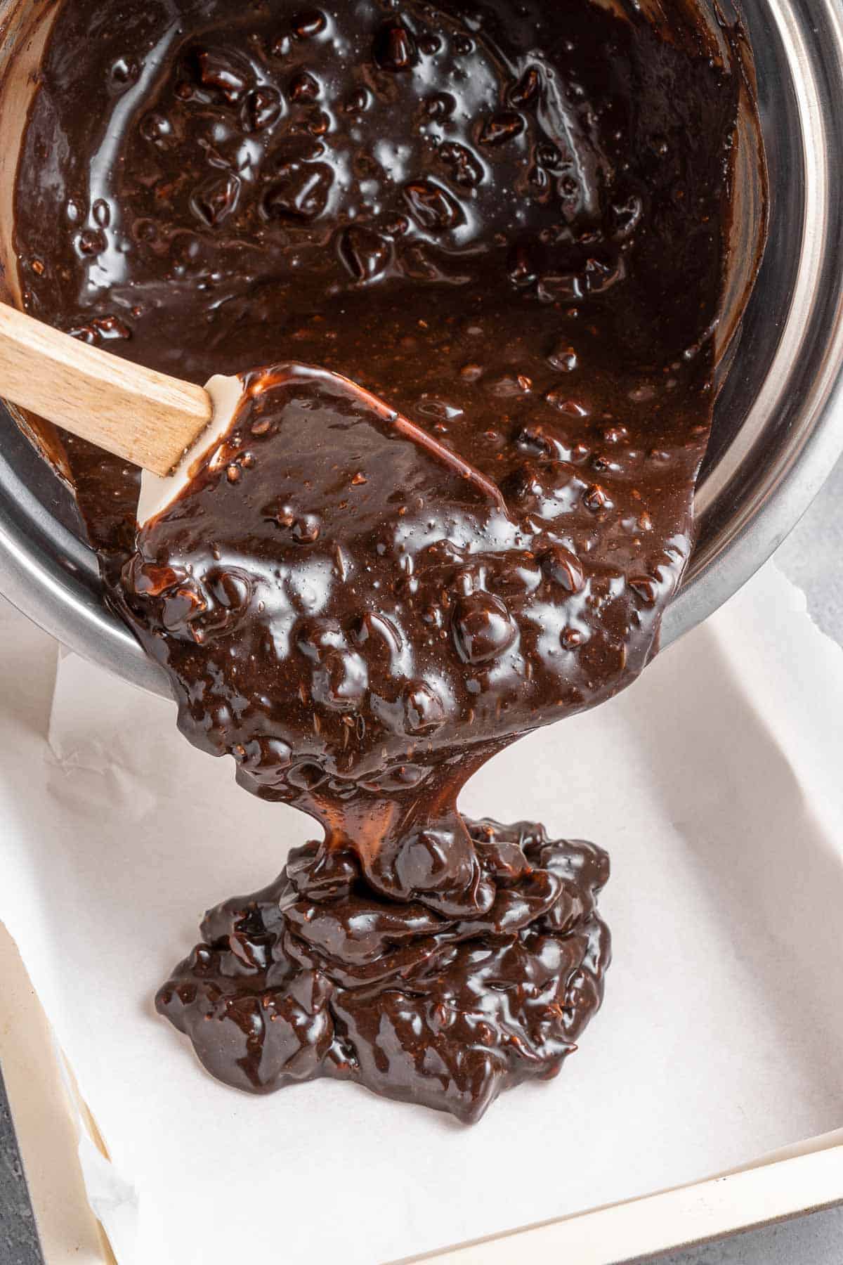 Chocolate fudge batter being poured into a square pan with a wooden spoon.