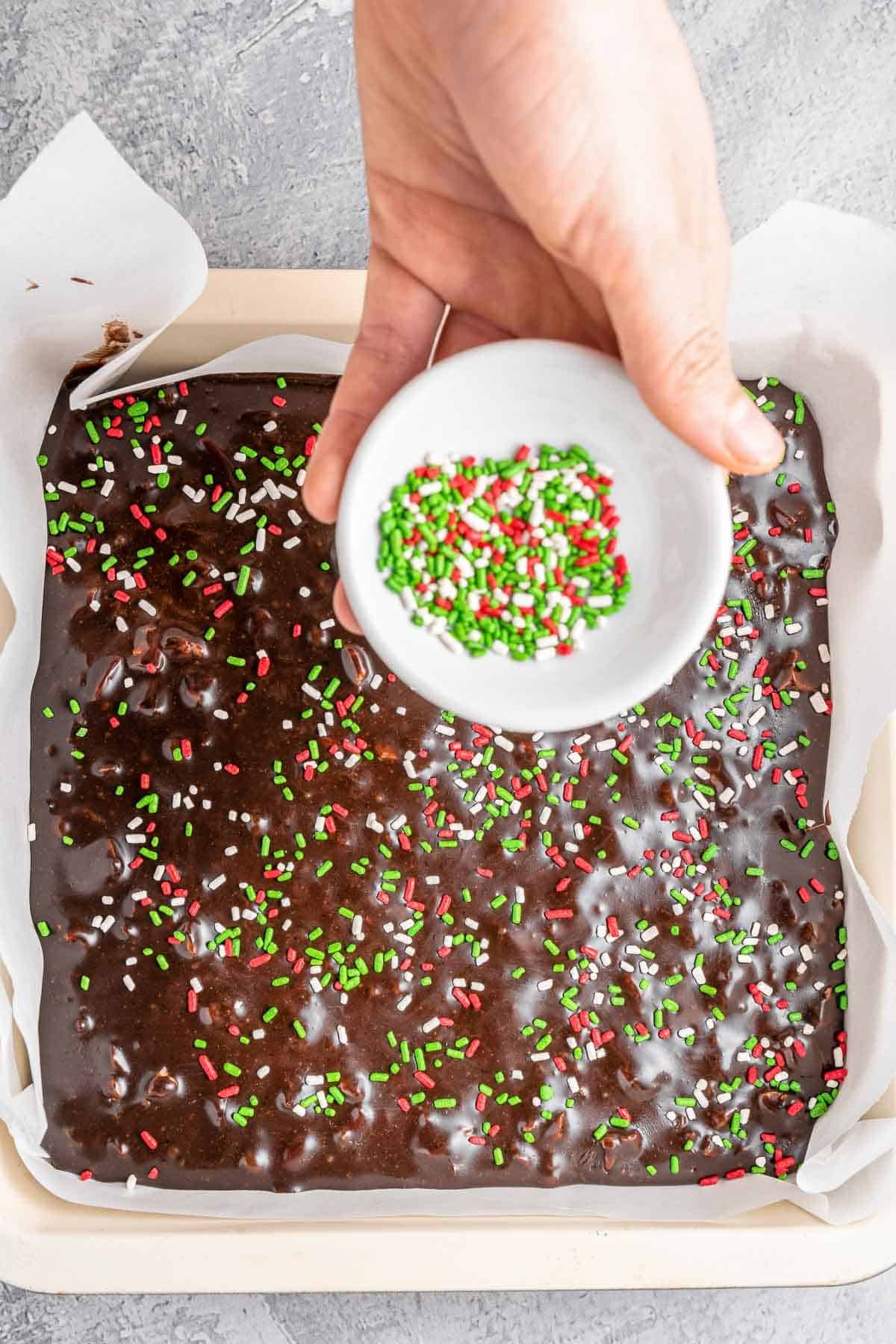 A hand sprinkling holiday colored sprinkles over chocolate fudge in a square baking dish.