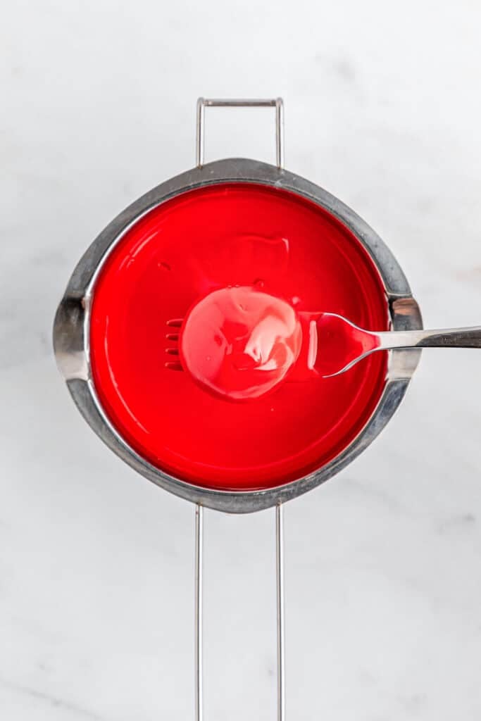 A fork holding a red dipped ball over a pot of red melted candy wafers.