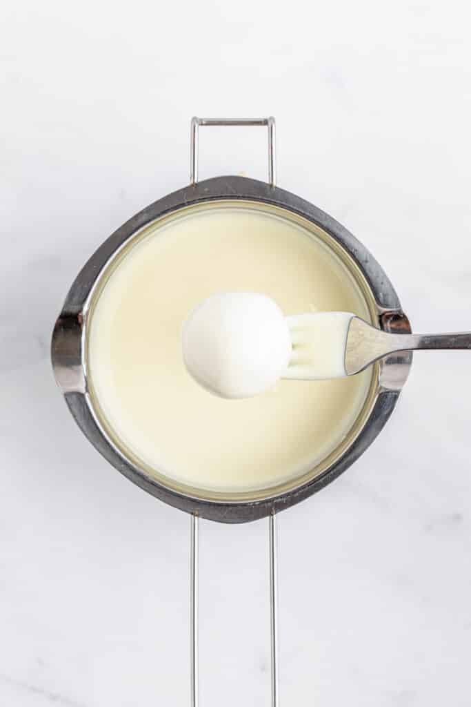 A fork holding a white dipped ball over a pot of white melted candy wafers.