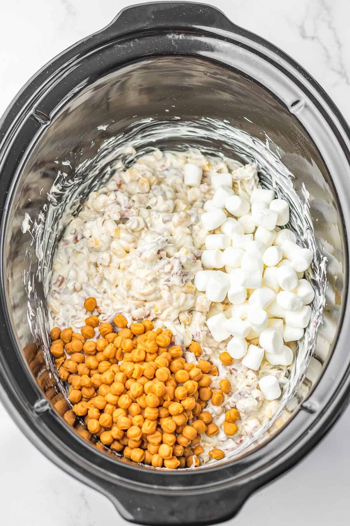 A crock pot filled with a white almond bark, pretzel and peanut mixture with mini marshmallows and caramel bits added in.
