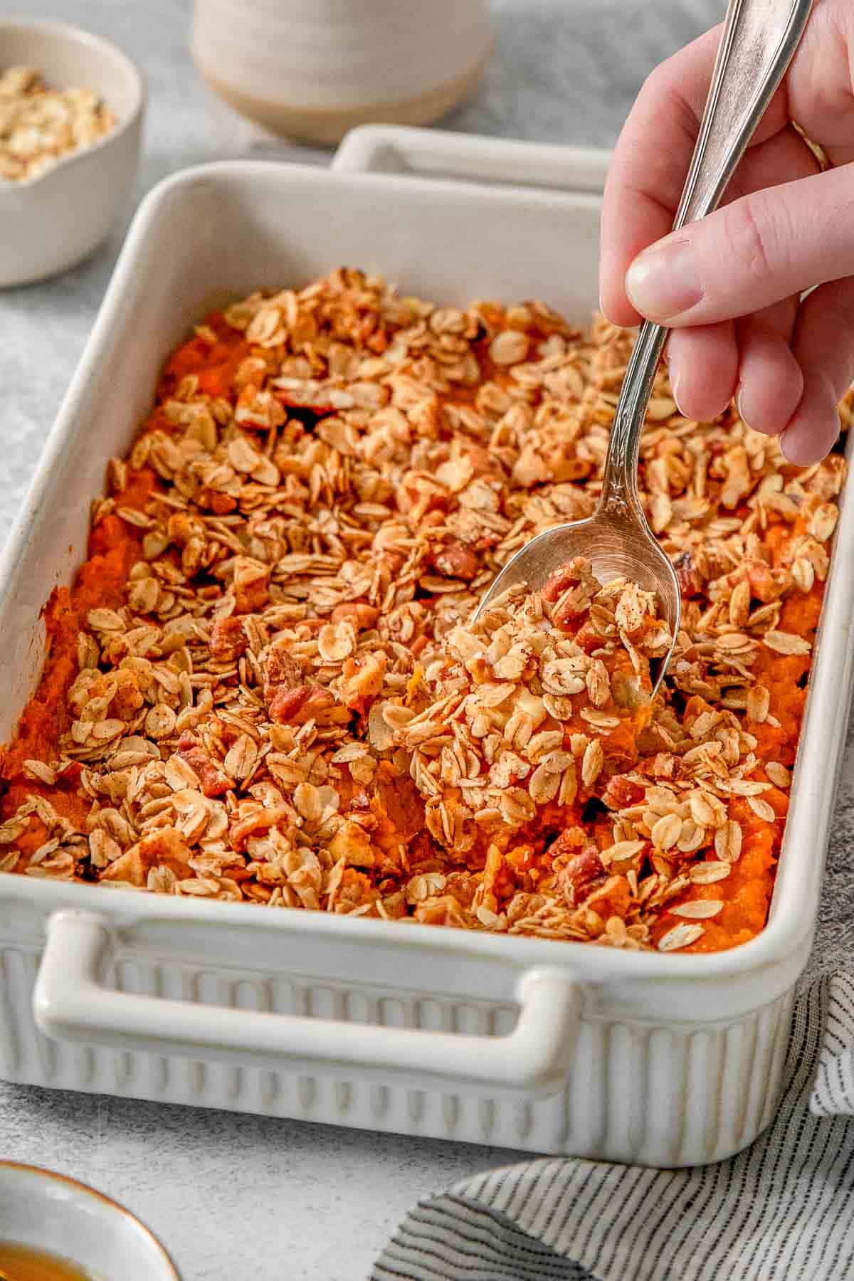 Woman's hand scooping sweet potato casserole out of a baking dish with a silver spoon.