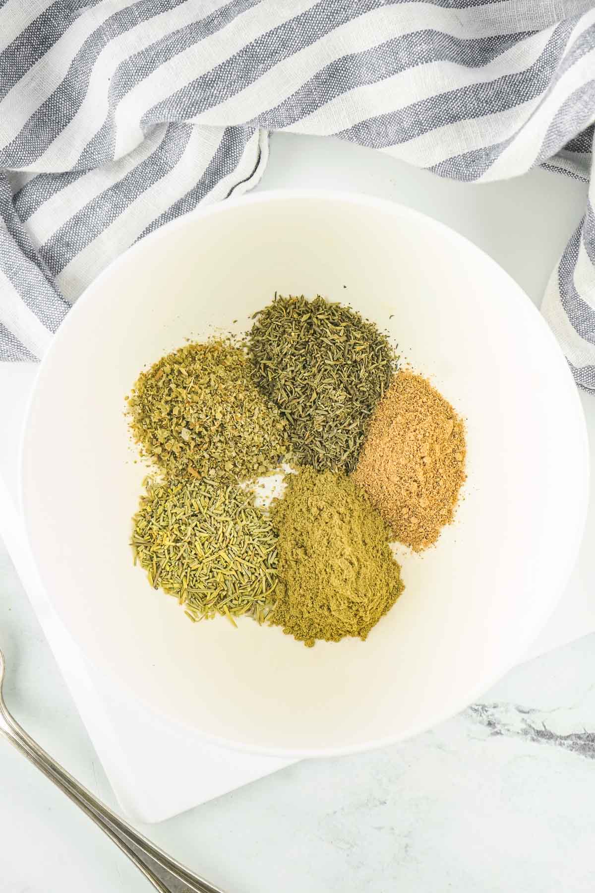 five spices for an easy homemade poultry seasoning recipe in piles in a white bowl.