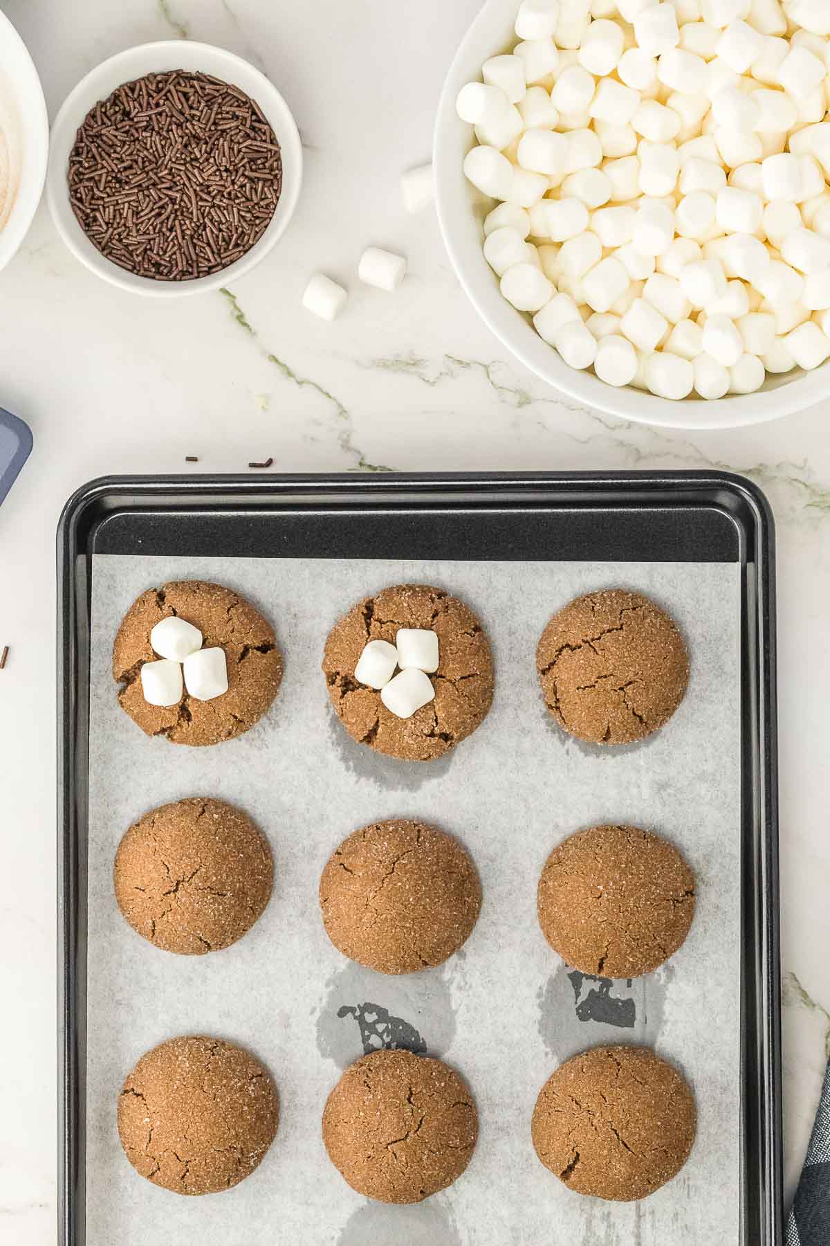 Baked hot chocolate cookies on a baking sheet being topped with mini marshmallows.