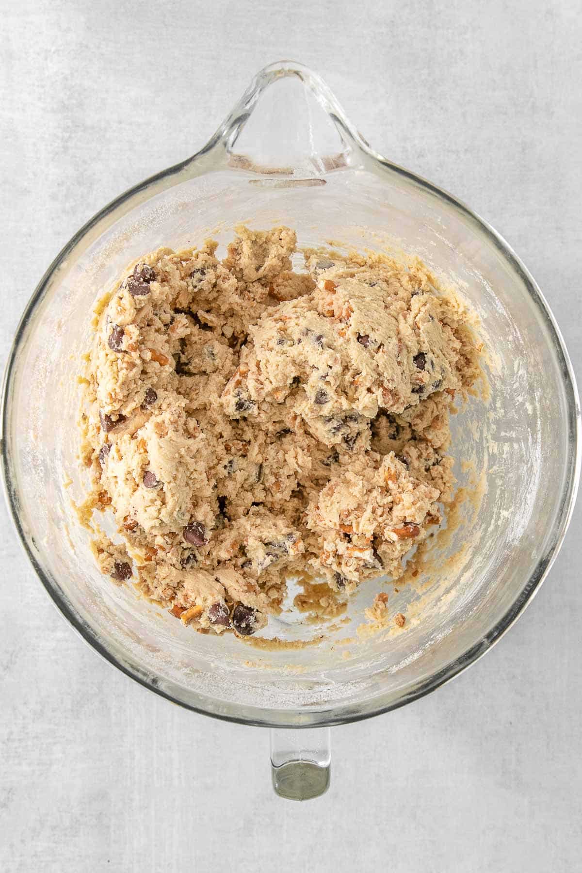 Chocolate chips and pretzels added to cookie dough in a glass bowl.