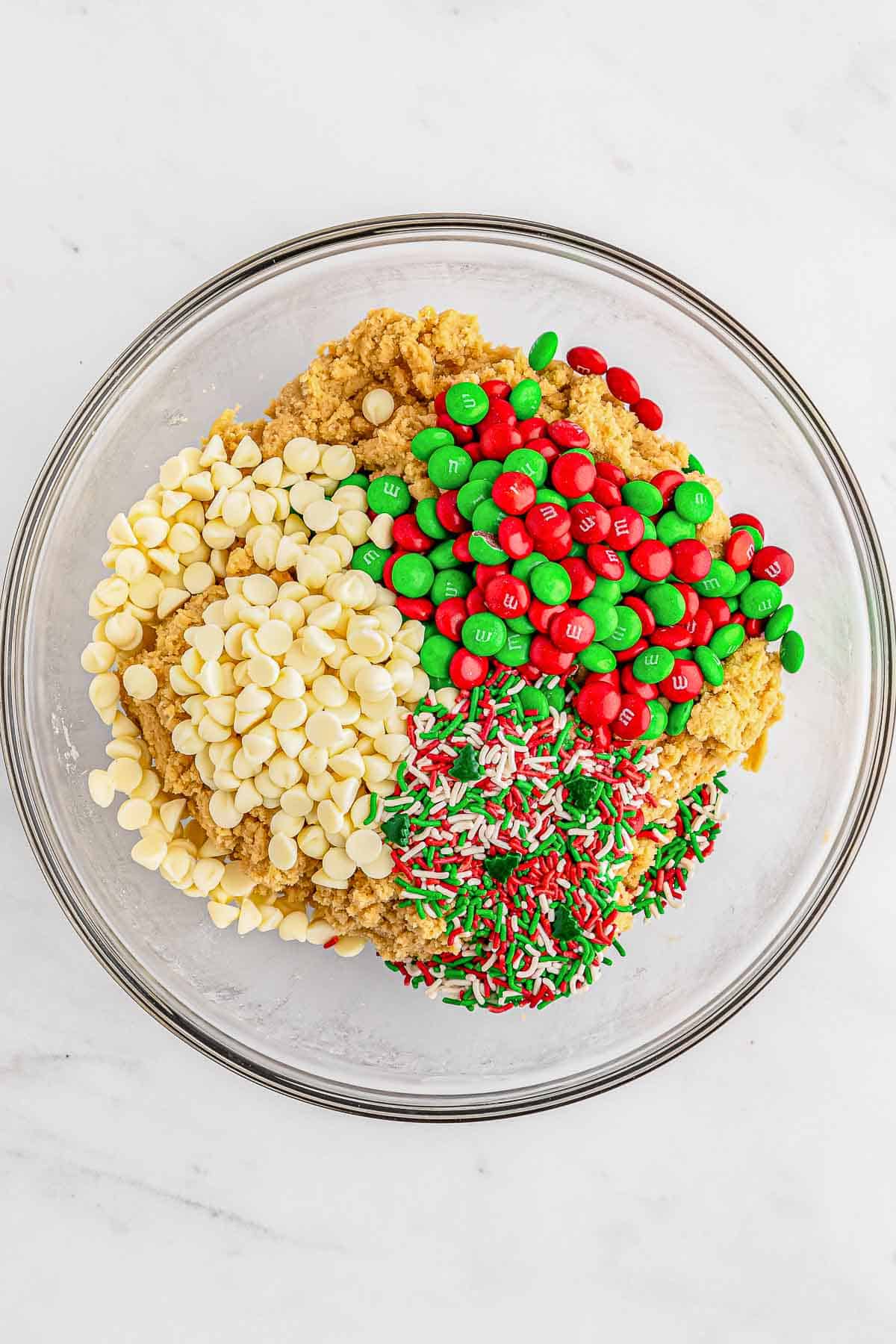 A glass bowl filled with cookie dough ingredients with m&m's, white chocolate chips and sprinkles.