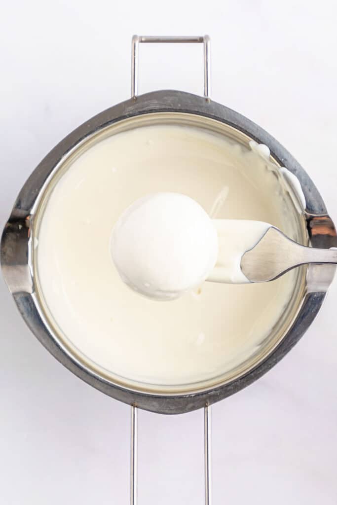 A fork is being used to dip a ball of cookie dough into white candy melt.