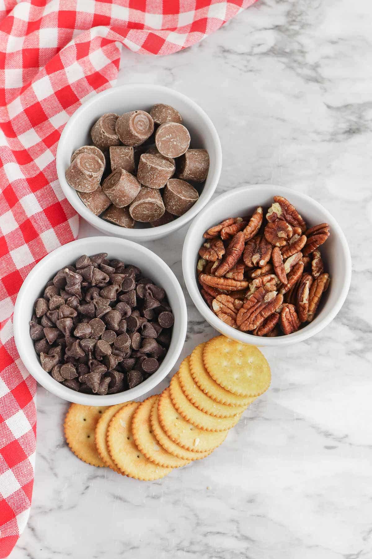 Three bowls of chocolate chips, pecans and chocolate chips and several ritz crackers on a marble table.