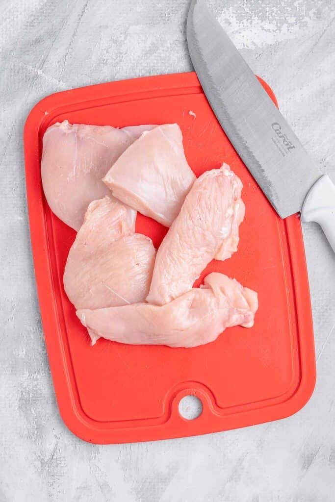 A red cutting board with thin raw chicken pieces on it and a knife resting on the side.