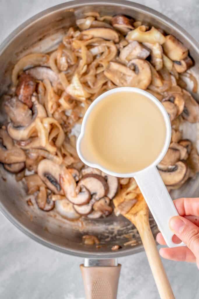 A hand pouring a white wine over sautéed mushrooms in a pan.