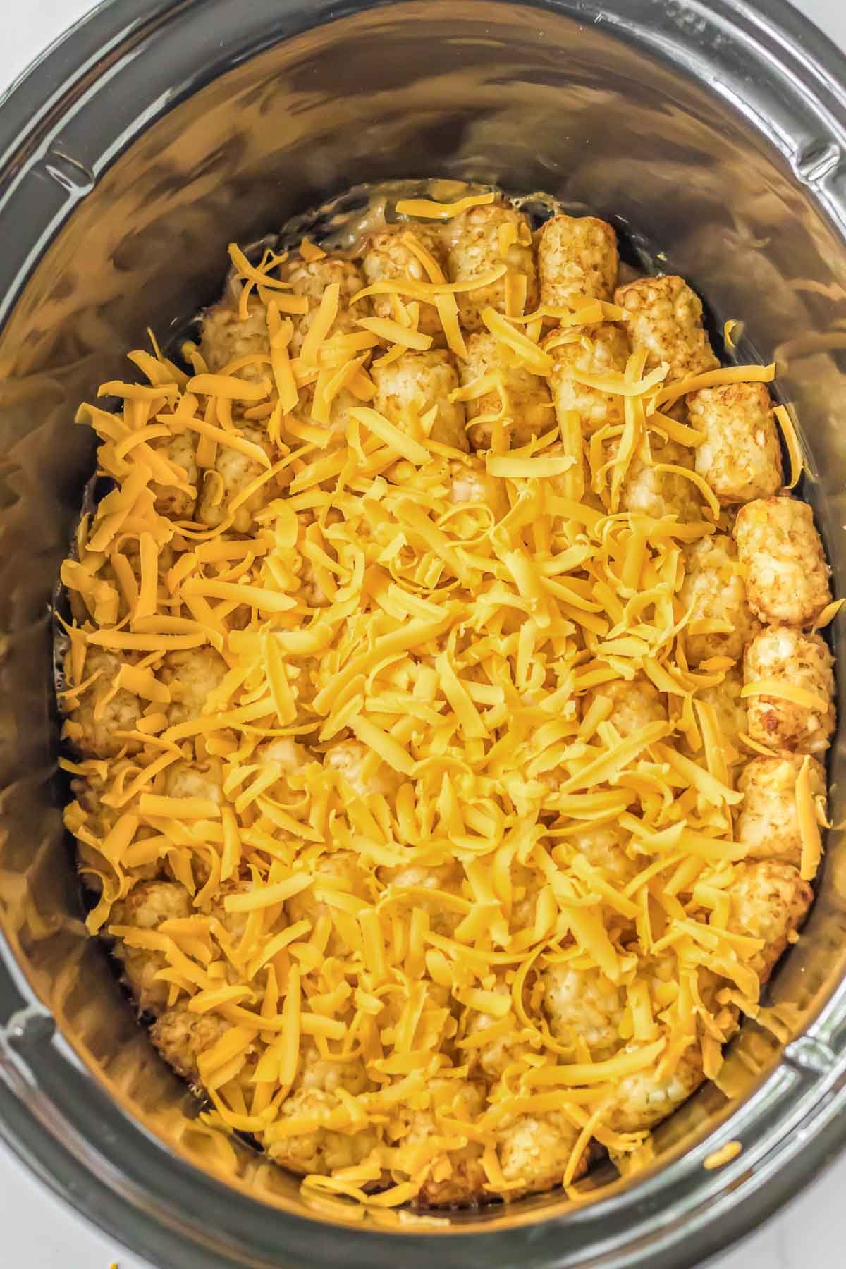 A crock pot full of tater tots topped with shredded cheese.