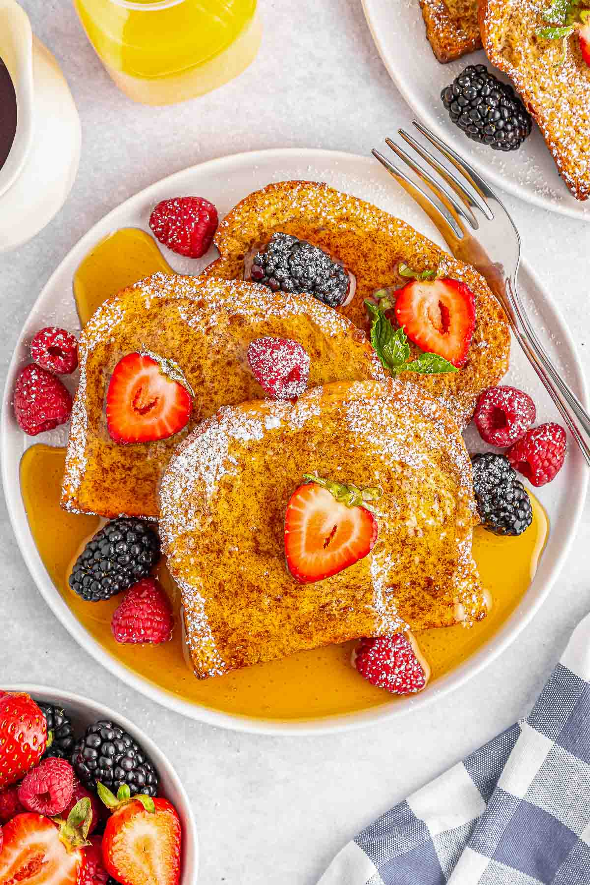 A white plate of air fryer french toast topped with maple syrup, powdered sugar, strawberries and raspberries .
