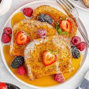 A white plate of air fryer french toast topped with maple syrup, powdered sugar, strawberries and raspberries .