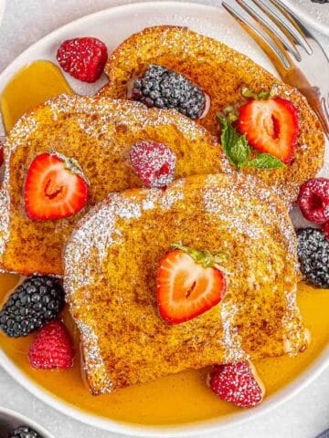 A white plate of air fryer french toast topped with maple syrup, powdered sugar, strawberries and raspberries .