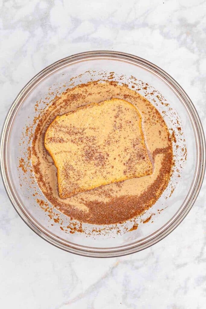 A slice of bread in a glass mixing bowl coated in the egg mixture.