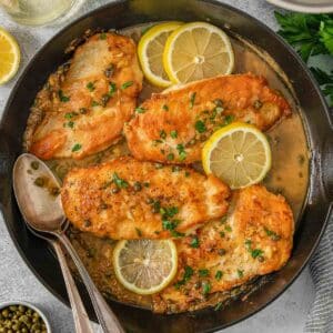Chicken picata coated in sauce in a skillet topped with lemon and parsley.