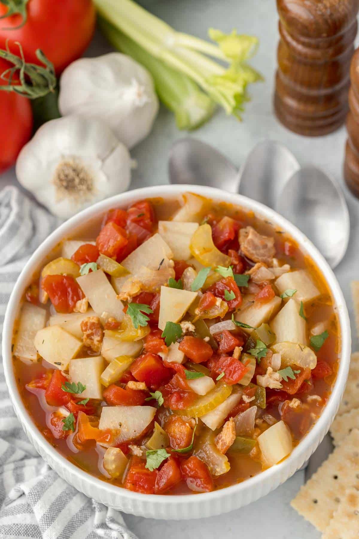 A white bowl of manhattan clam chowder recipe with crackers and three silver spoons beside it.