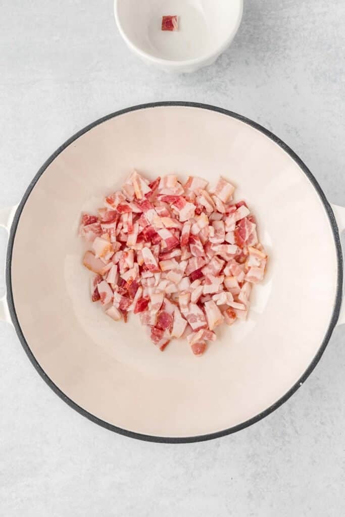 A white dutch oven filled with chopped raw bacon and a bowl next to it.