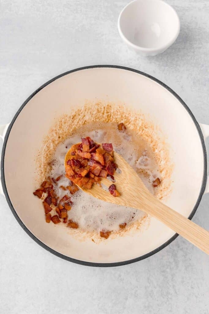 chopped bacon being scooped out of a white pot with a wooden spoon.