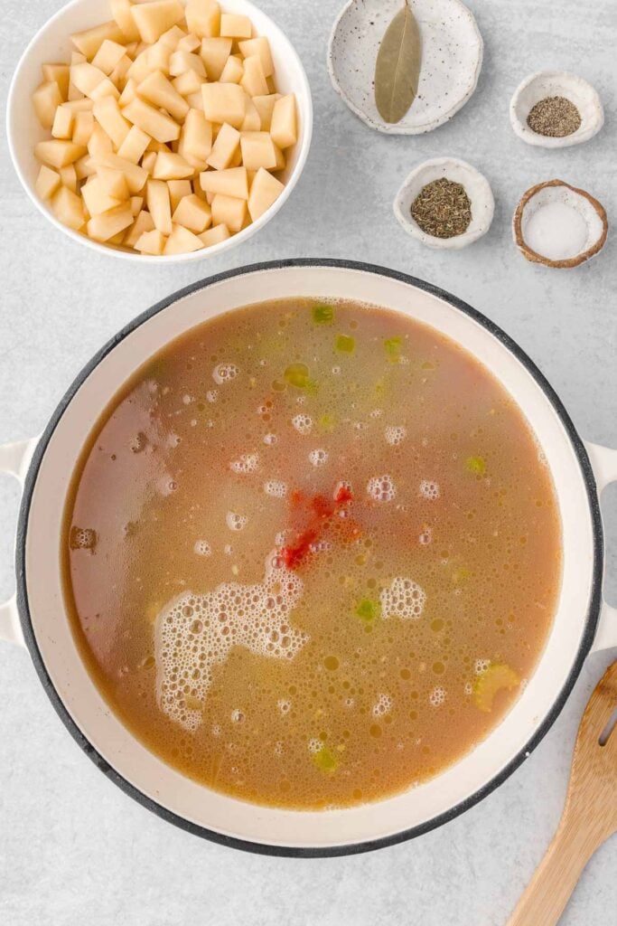 A white pot of broth and diced tomatoes with a bowl of diced potatoes beside it.