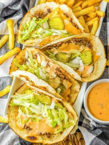 Smash burger tacos topped with lettuce, pickles and onion on a plate with french fries.
