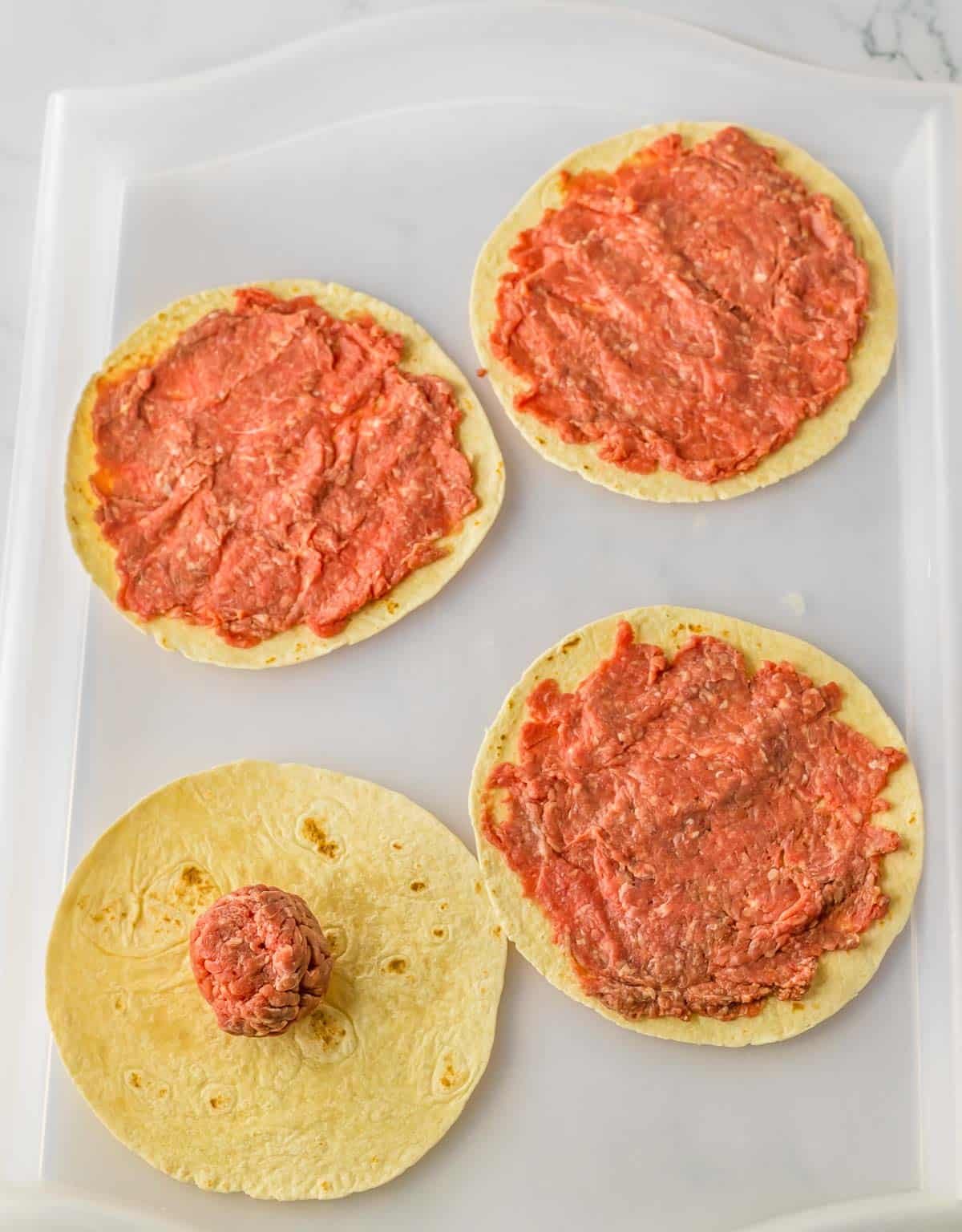 Four tortillas with meat scooped and flattened on a baking sheet.