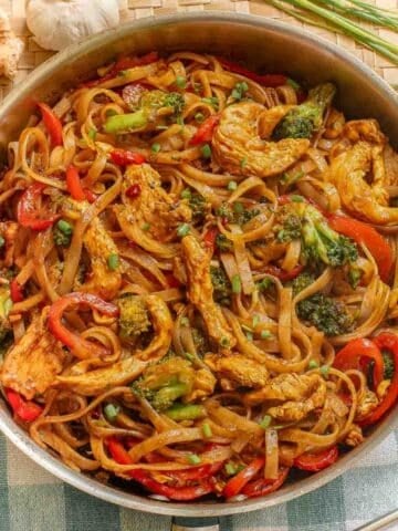 A skillet full of a drunken noodles recipe with rice noodles, chicken and red bell peppers and broccoli.