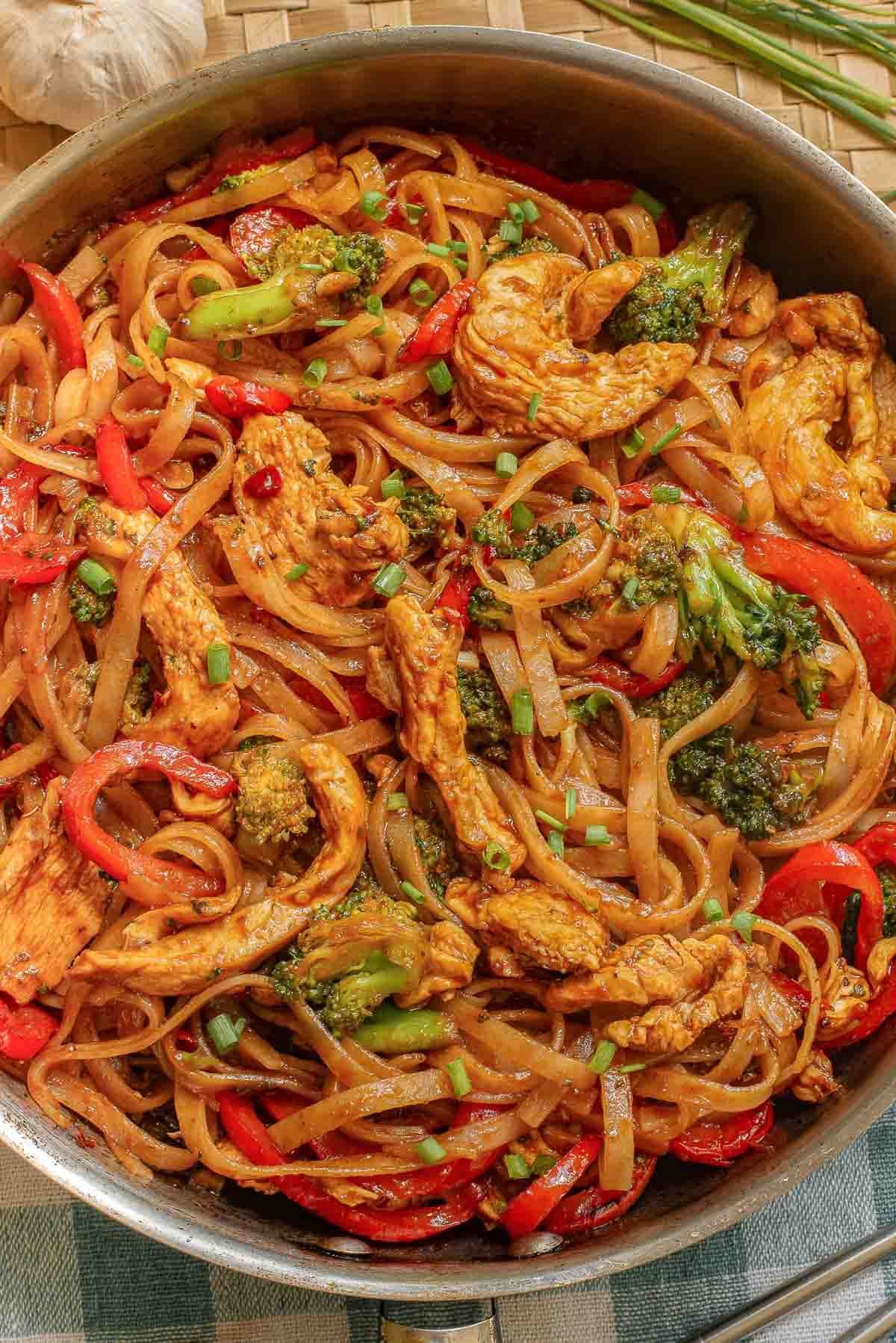 A pan full of rice noodles, chicken, red bell peppers, and broccoli in an Asian sauce.