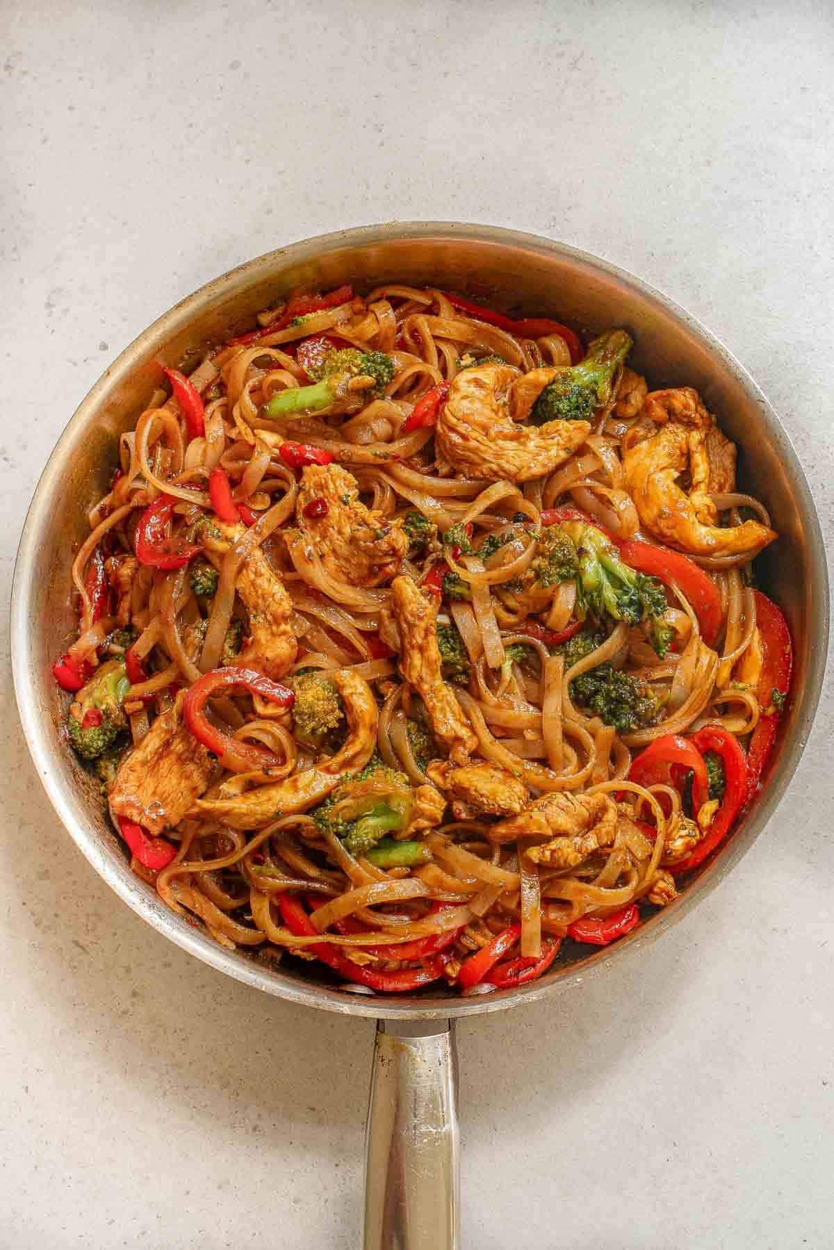 drunken noodles recipe cooked in a silver skillet with rice noodles, chicken and red bell peppers and broccoli in a Asian sauce.