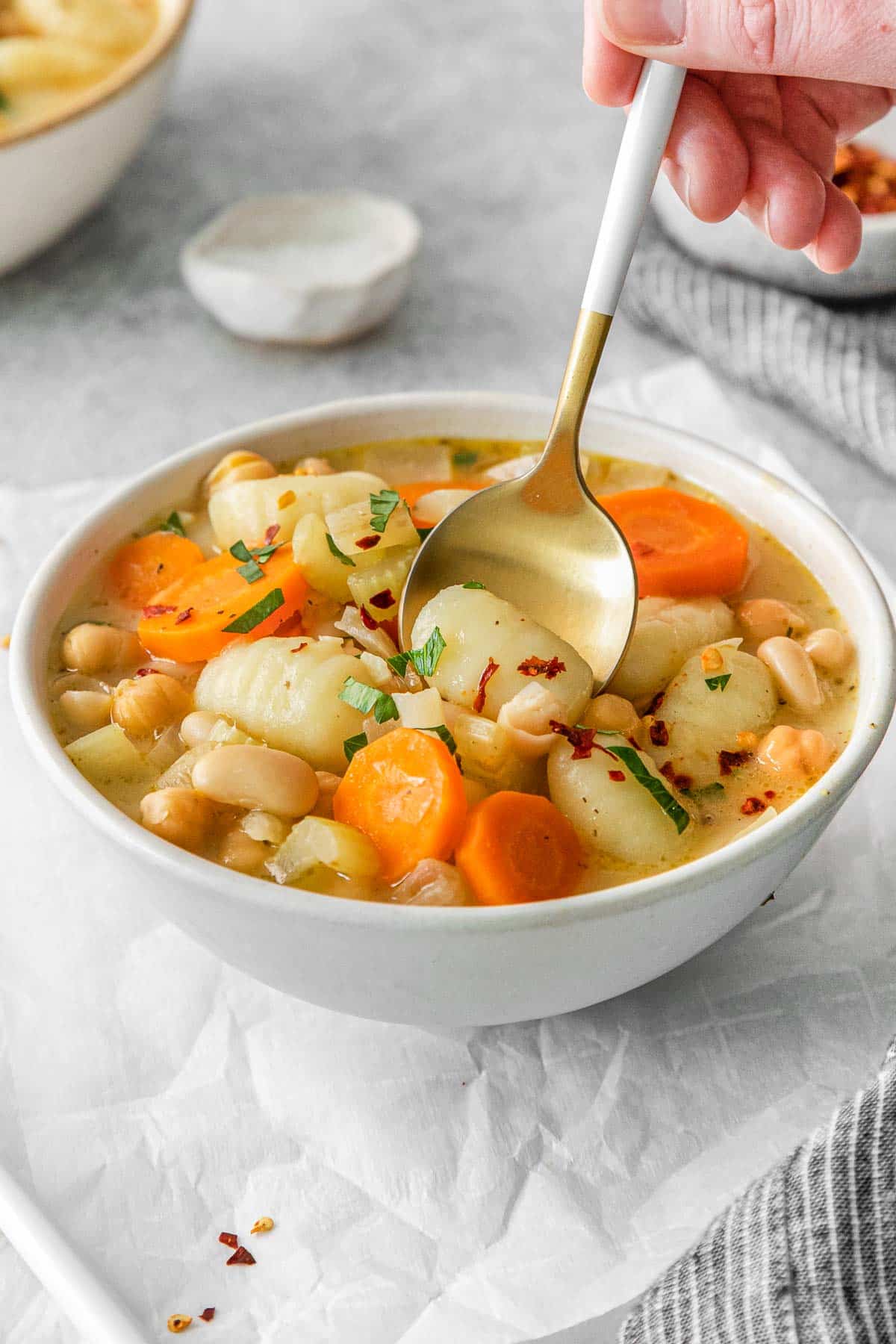 A person holding a spoonful of gnocchi soup with carrots and beans in a white bowl.